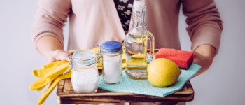 Photo d'une personne tenant un plateau avec différents produits nécessaires à la fabrication de produits ménagers : du citron, du bicarbonate de soude et du vinaigre ainsi que des éponges et une bouteille